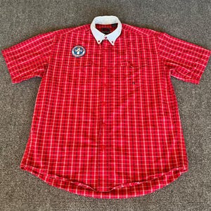 May include: Red plaid short-sleeve shirt with a white collar. The shirt has a button-down front and a small patch on the left chest. The plaid pattern is red, black, and white.