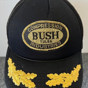 May include: Black trucker hat with a gold embroidered oval patch that says "Compression Bush Tulsa Industries". The hat also has two gold embroidered leaf designs on the front.