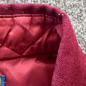 May include: A maroon jacket featuring a textured knit collar and a quilted interior lining. The garment includes a small blue fabric tag attached to the inner seam, showcasing a consistent deep red color palette throughout the visible sections.