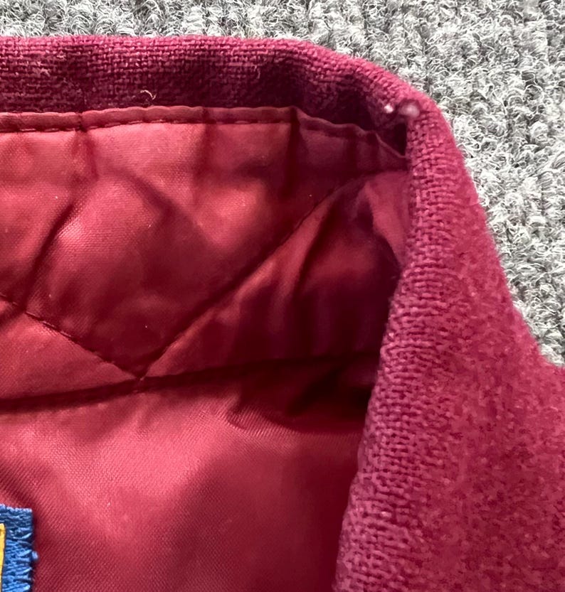 May include: A maroon jacket featuring a textured knit collar and a quilted interior lining. The garment includes a small blue fabric tag attached to the inner seam, showcasing a consistent deep red color palette throughout the visible sections.