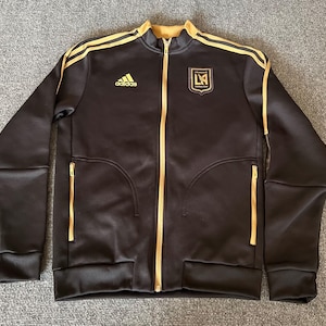 May include: Black zip-up jacket with gold accents, featuring the Adidas logo and a team crest. The jacket has gold stripes on the shoulders and gold zippers on the pockets and front. The jacket is a sporty style.