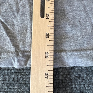 Pu&ograve; includere: Primo piano di un tessuto grigio con un righello di legno posizionato verticalmente a destra. Il righello mostra misure da 58 a 71 cm. Il tessuto sembra provenire da una maglietta o da un capo di abbigliamento simile.