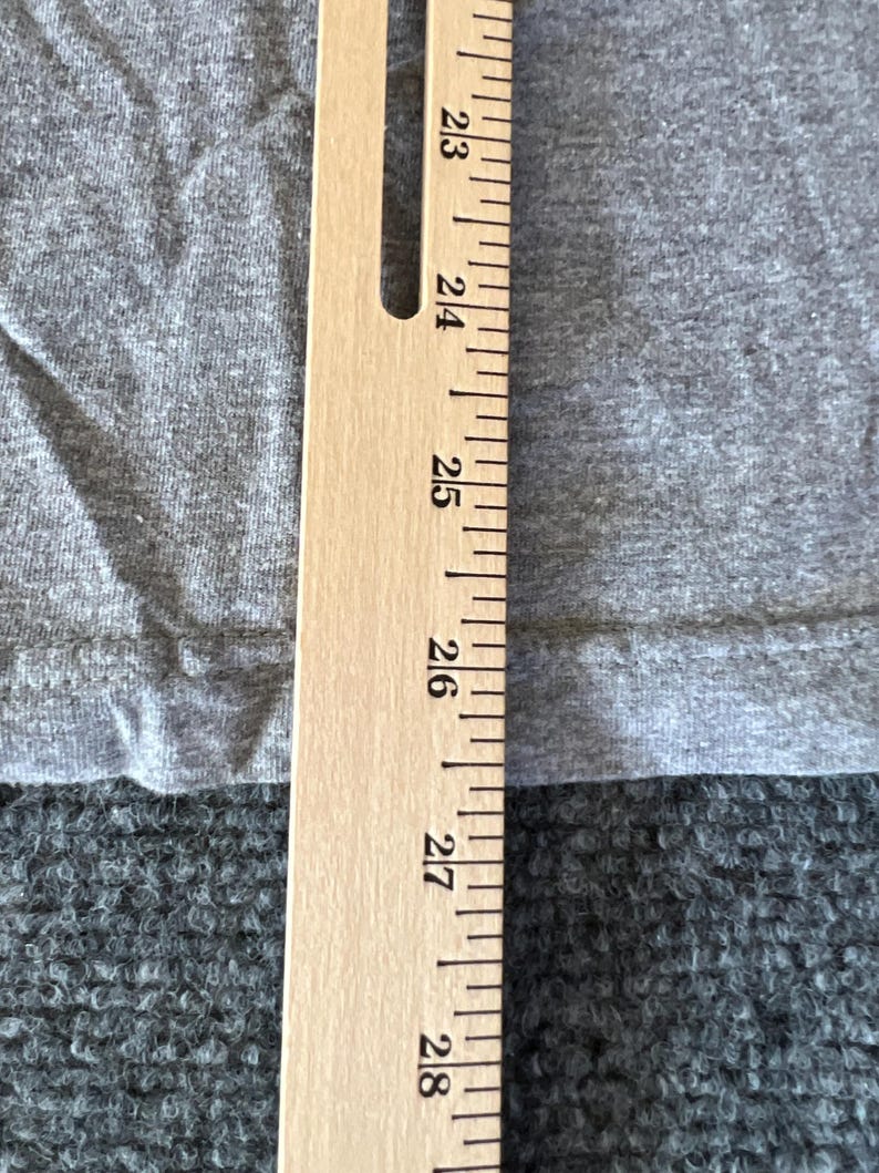 Pu&ograve; includere: Primo piano di un tessuto grigio con un righello di legno posizionato verticalmente a destra. Il righello mostra misure da 58 a 71 cm. Il tessuto sembra provenire da una maglietta o da un capo di abbigliamento simile.