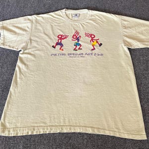 May include: Cream-colored t-shirt featuring a colorful graphic of three stylized figures dancing. The text "METRIS SPRING ART FAIR" and "AUGUST 13, 2003" are printed below the figures. The shirt has short sleeves and a crew neck.