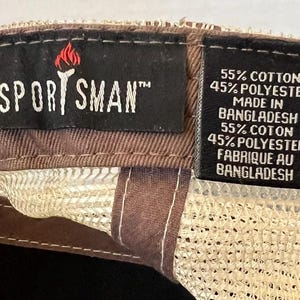 Op de afbeelding: Close-up van een bruine en beige baseballcap. De cap heeft een mesh achterkant en een bruine klep. Het label leest "SPORTSMAN" met een fakkel logo. Het label geeft ook aan dat de cap is gemaakt van 55% katoen en 45% polyester.