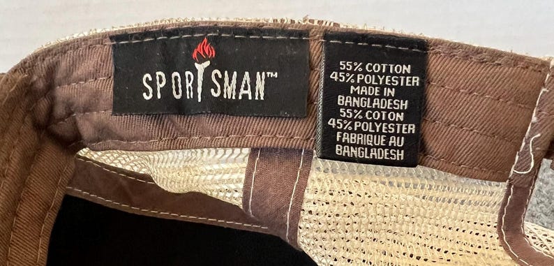 Op de afbeelding: Close-up van een bruine en beige baseballcap. De cap heeft een mesh achterkant en een bruine klep. Het label leest "SPORTSMAN" met een fakkel logo. Het label geeft ook aan dat de cap is gemaakt van 55% katoen en 45% polyester.