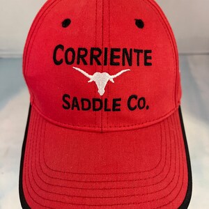 May include: Red baseball cap with black stitching and a black brim. The front of the cap features a white embroidered longhorn steer head with the text "Corriente Saddle Co."