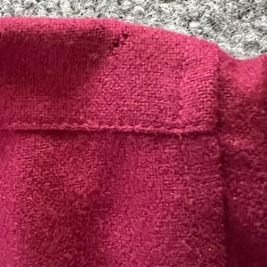 May include: A close-up view of a deep magenta or dark pink garment featuring a textured, soft fabric surface. The item displays a horizontal seam line across the upper section with visible stitching detail and a small, singular snag in the fabric.