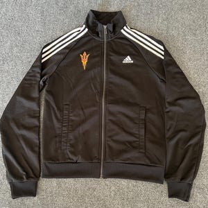 May include: Black Adidas track jacket with white stripes on the shoulders and sleeves. Features a full-length zipper, two front pockets, and the Adidas logo on the chest. The Arizona State University logo is on the left chest.