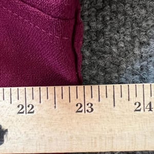 May include: A deep magenta fabric garment positioned against a wooden ruler. The ruler shows measurements from 22 inches to 24 inches, clearly indicating the scale of the textile edge and the visible stitching along the seam of the magenta material.