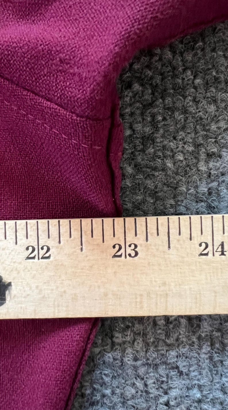 May include: A deep magenta fabric garment positioned against a wooden ruler. The ruler shows measurements from 22 inches to 24 inches, clearly indicating the scale of the textile edge and the visible stitching along the seam of the magenta material.
