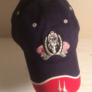 May include: A navy blue baseball cap with a white and pink embroidered logo featuring a horseshoe, a rose, and a horse's head. The cap has a red brim with a white trim and a white star logo.