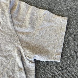 Pu&ograve; includere: Primo piano di una maglietta grigia a maniche corte. Il tessuto ha una trama m&eacute;lange, con cuciture visibili sulla spalla e sulla manica. La manica &egrave; ripiegata sull'orlo.