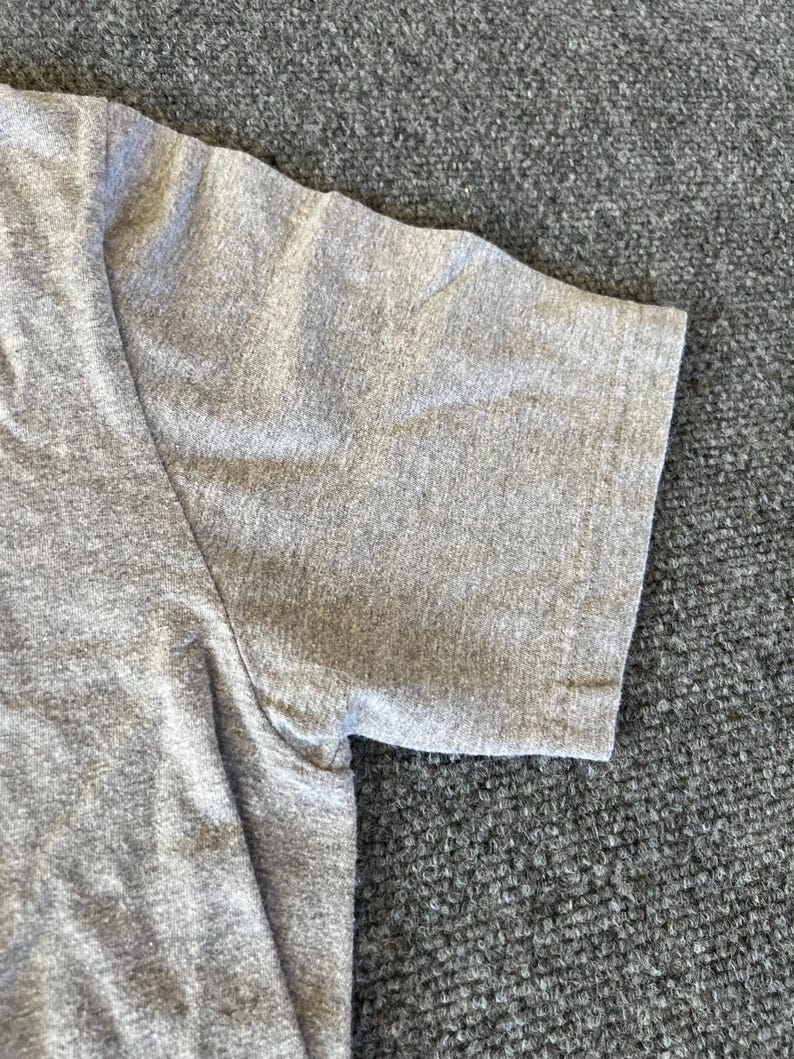 Pu&ograve; includere: Primo piano di una maglietta grigia a maniche corte. Il tessuto ha una trama m&eacute;lange, con cuciture visibili sulla spalla e sulla manica. La manica &egrave; ripiegata sull'orlo.