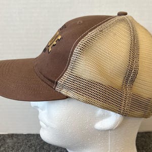 Op de afbeelding: Een bruine baseballcap met een beige mesh achterkant. De cap heeft een gebogen klep en een goudkleurig logo op de voorkant. De cap staat op een witte mannequinhoofd.