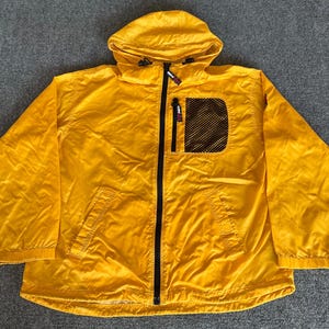 May include: A bright yellow hooded jacket with a full-length black zipper. The jacket has a mesh pocket on the chest and a small logo near the zipper. The jacket is made of a lightweight material.