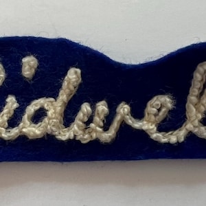 May include: A dark blue felt patch featuring the name Sidwell embroidered in white thread. The lettering is in a cursive script style, and the patch has an irregular, rounded rectangular shape with a soft, fuzzy texture on the blue fabric.