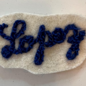 May include: A white felt patch featuring the word "Lopey" embroidered in a cursive script using thick, textured royal blue yarn. The patch has an irregular, rounded rectangular shape with a soft, fuzzy surface texture throughout the material.
