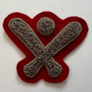May include: A red felt patch featuring a textured, light brown embroidered design of two crossed baseball bats positioned beneath a small, round baseball. The patch has a scalloped edge and a raised, tactile surface created by the embroidery.