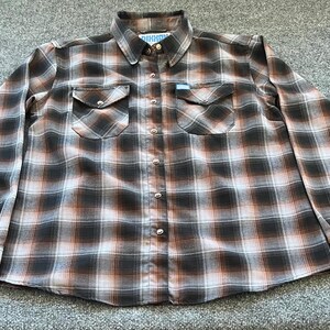 May include: A long-sleeved, button-down flannel shirt in a brown, black, and white plaid pattern. The shirt has two chest pockets with button closures and buttoned cuffs. The collar has a button closure. The shirt is laid flat on a gray surface.
