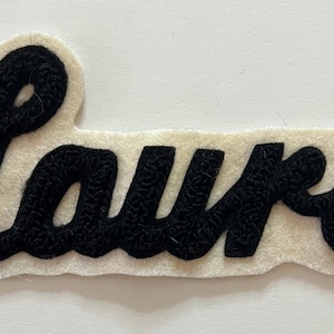 May include: A black embroidered patch featuring the name Laura in a cursive script font. The black lettering is stitched onto a cream-colored felt backing that follows the contour of the letters, creating a distinct border around the text.