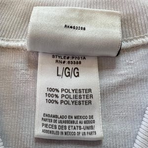 Op de afbeelding: Close-up van een wit kledinglabel met de tekst "RN#63388" en "STYLE#:P701A RN#:63388 L/G/G". Het label vermeldt ook "100% POLYESTER" en tekst in het Spaans en Frans over assemblage in Mexico.