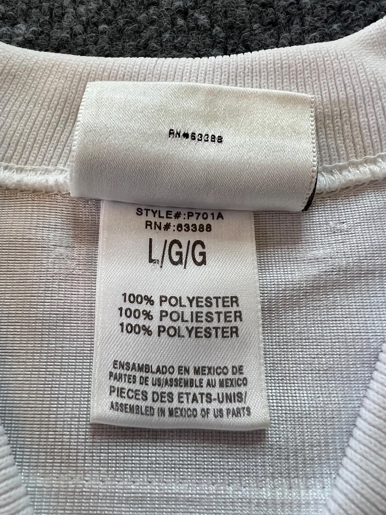 Op de afbeelding: Close-up van een wit kledinglabel met de tekst "RN#63388" en "STYLE#:P701A RN#:63388 L/G/G". Het label vermeldt ook "100% POLYESTER" en tekst in het Spaans en Frans over assemblage in Mexico.