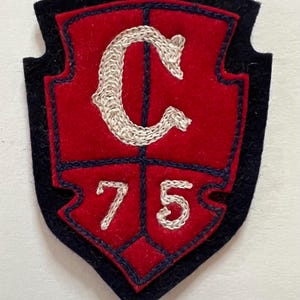 May include: A shield-shaped felt patch featuring a red center with a dark blue border. The patch displays an embroidered white letter C above the number 75, with dark blue stitching dividing the red sections into four distinct quadrants.