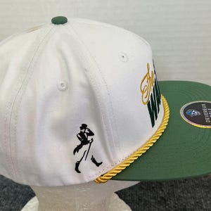 May include: White and green golf cap with the text "The Way" in gold and green. The cap features a black silhouette of a man walking with a cane. A gold rope trim separates the white crown from the green brim.