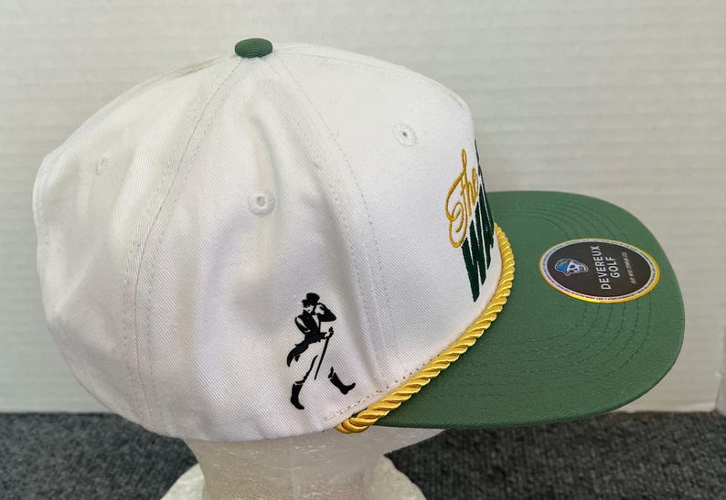 May include: White and green golf cap with the text "The Way" in gold and green. The cap features a black silhouette of a man walking with a cane. A gold rope trim separates the white crown from the green brim.
