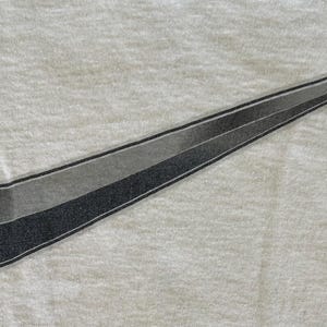 May include: A white t-shirt featuring a large, stylized Nike swoosh logo. The swoosh is a gradient of gray and black, with a textured appearance. The shirt's fabric has a subtle, textured pattern.