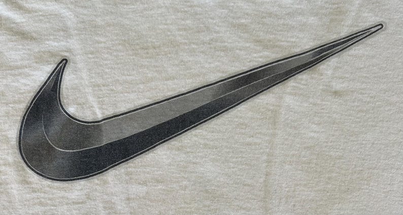 May include: A white t-shirt featuring a large, stylized Nike swoosh logo. The swoosh is a gradient of gray and black, with a textured appearance. The shirt's fabric has a subtle, textured pattern.