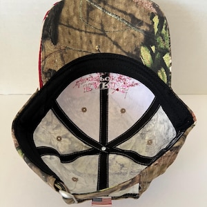 May include: Camouflage baseball cap with a curved brim. The cap features a brown, green, and tan pattern on the exterior and a white interior with text. A small American flag tag is sewn on the back.