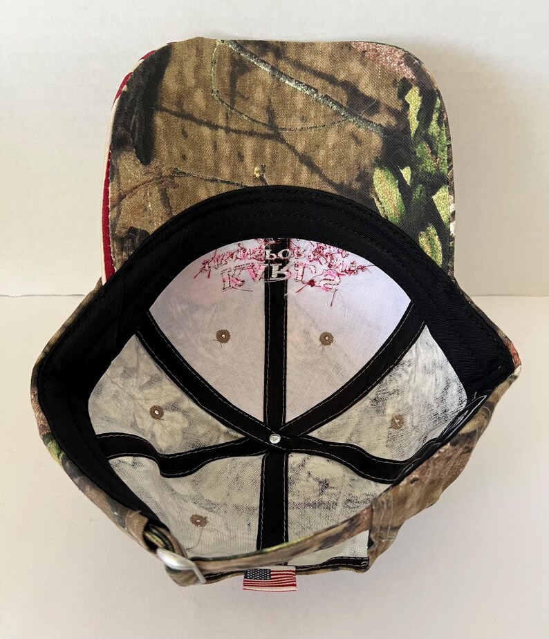 May include: Camouflage baseball cap with a curved brim. The cap features a brown, green, and tan pattern on the exterior and a white interior with text. A small American flag tag is sewn on the back.