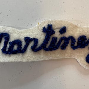 May include: A white felt patch featuring the name Martinez embroidered in dark blue thread. The lettering is in a cursive script style, and the white felt backing is cut to follow the shape of the embroidered name in a rectangular format.
