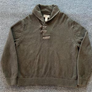 May include: An olive green long-sleeve sweater featuring a thick ribbed knit texture, a wide shawl collar, and a front placket with three toggle-style closures. A Duluth Trading Co. brand label is visible on the inside of the collar neckline.