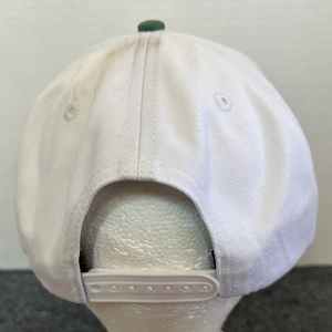 May include: A white baseball cap with a green button on top and a white adjustable snap closure. The cap is made of a sturdy fabric and has a curved brim. The back of the cap is visible.