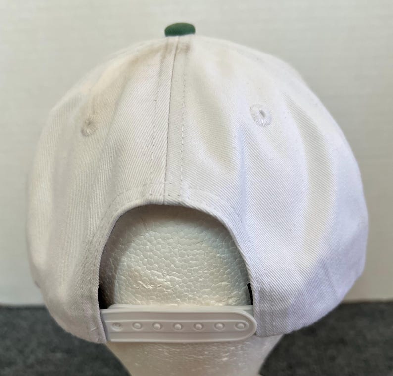 May include: A white baseball cap with a green button on top and a white adjustable snap closure. The cap is made of a sturdy fabric and has a curved brim. The back of the cap is visible.