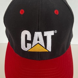 May include: Black and red baseball cap with a white CAT logo and a yellow triangle. The logo is embroidered on the front of the cap.