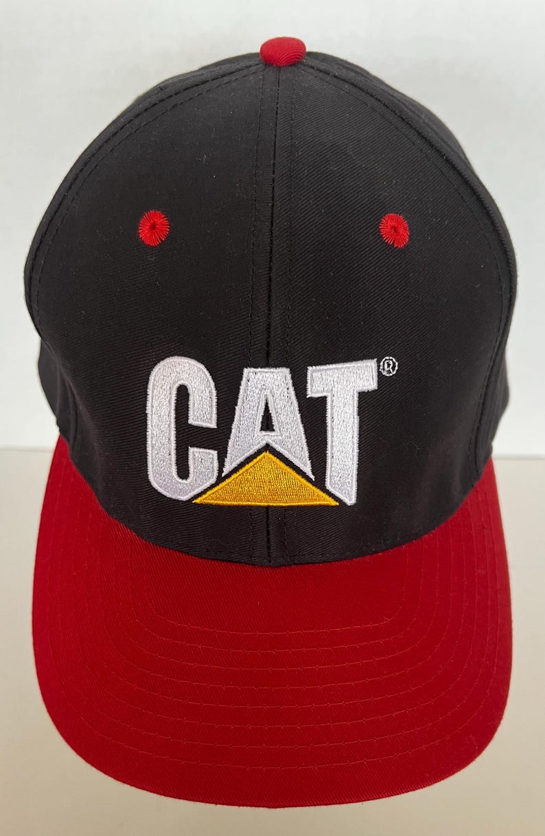 May include: Black and red baseball cap with a white CAT logo and a yellow triangle. The logo is embroidered on the front of the cap.