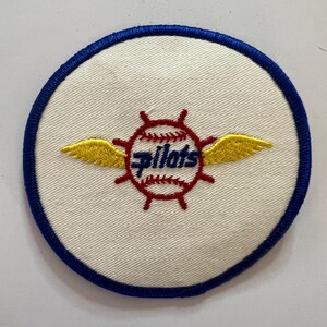May include: A circular embroidered patch with a white background and a blue border. The center features a red ship's wheel design overlaid on a baseball, with yellow wings extending from the sides and the word "pilots" in blue script text.