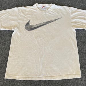 May include: Off-white short-sleeved t-shirt featuring a large, gradient gray Nike swoosh logo across the chest. The shirt has a crew neck and a small Nike logo tag at the neckline. The fabric appears slightly worn.