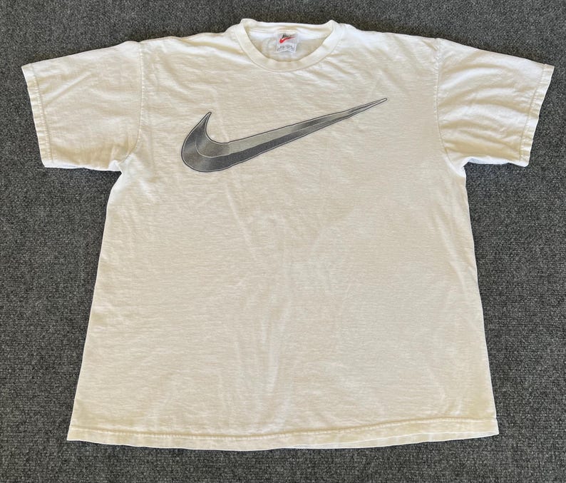 May include: Off-white short-sleeved t-shirt featuring a large, gradient gray Nike swoosh logo across the chest. The shirt has a crew neck and a small Nike logo tag at the neckline. The fabric appears slightly worn.