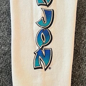 Peut inclure: Une manche de chemise &agrave; manches longues blanche avec le texte ROZON &eacute;crit verticalement. Les lettres pr&eacute;sentent un d&eacute;grad&eacute; de bleu clair &agrave; bleu fonc&eacute;, soulign&eacute;es de noir avec un symbole de marque d&eacute;pos&eacute;e &agrave; la fin du mot.