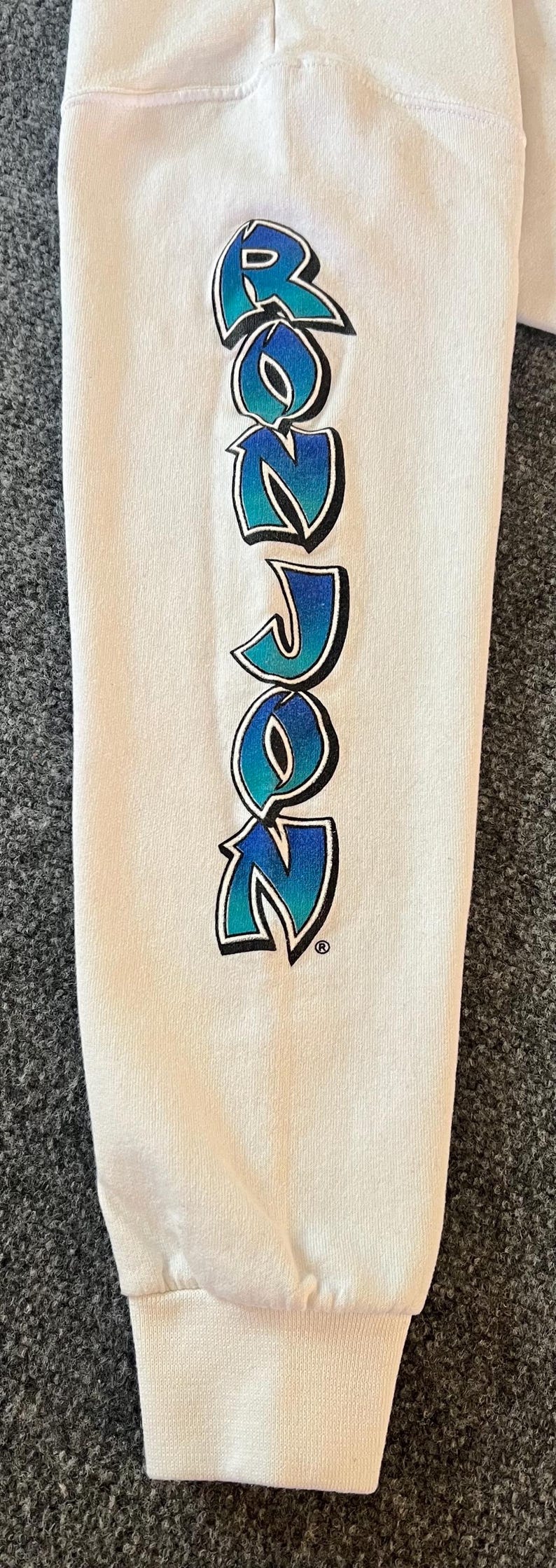 Peut inclure: Une manche de chemise &agrave; manches longues blanche avec le texte ROZON &eacute;crit verticalement. Les lettres pr&eacute;sentent un d&eacute;grad&eacute; de bleu clair &agrave; bleu fonc&eacute;, soulign&eacute;es de noir avec un symbole de marque d&eacute;pos&eacute;e &agrave; la fin du mot.