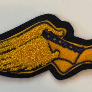 May include: A felt patch featuring a winged foot design in yellow and dark blue. The wing is rendered in a textured, raised yellow yarn embroidery, while the foot shape is made of smooth yellow felt with dark blue stitched outlines.