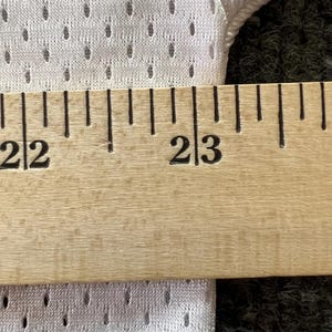 Op de afbeelding: Close-up van witte mesh stof met een liniaal die metingen weergeeft. De liniaal geeft een meting van 56 tot 58 cm aan. De stof heeft kleine, gelijkmatig verdeelde gaatjes.