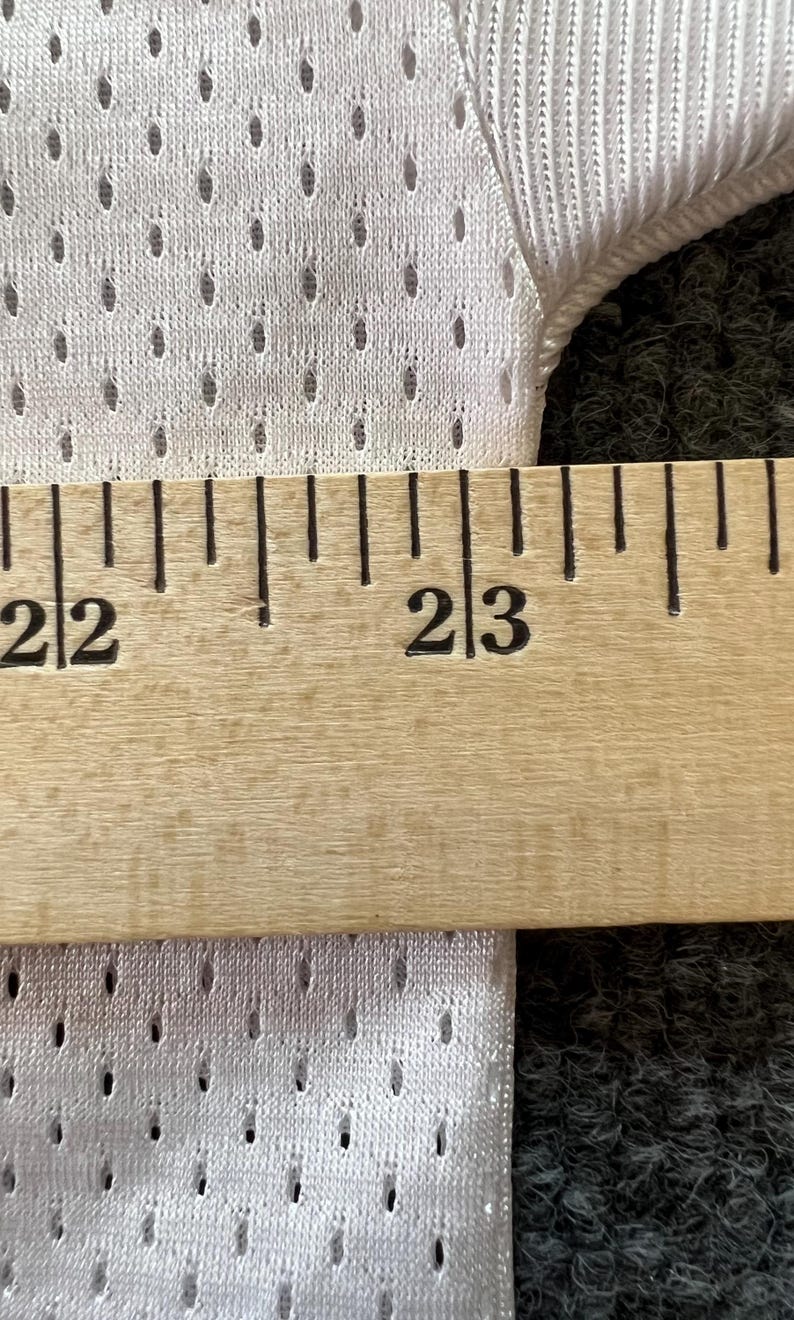 Op de afbeelding: Close-up van witte mesh stof met een liniaal die metingen weergeeft. De liniaal geeft een meting van 56 tot 58 cm aan. De stof heeft kleine, gelijkmatig verdeelde gaatjes.