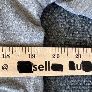 Pu&ograve; includere: Primo piano di un tessuto grigio con un righello di legno sopra. Il righello ha segni neri e la parola "sell" &egrave; parzialmente visibile. Il tessuto sembra essere un materiale a maglia morbido.