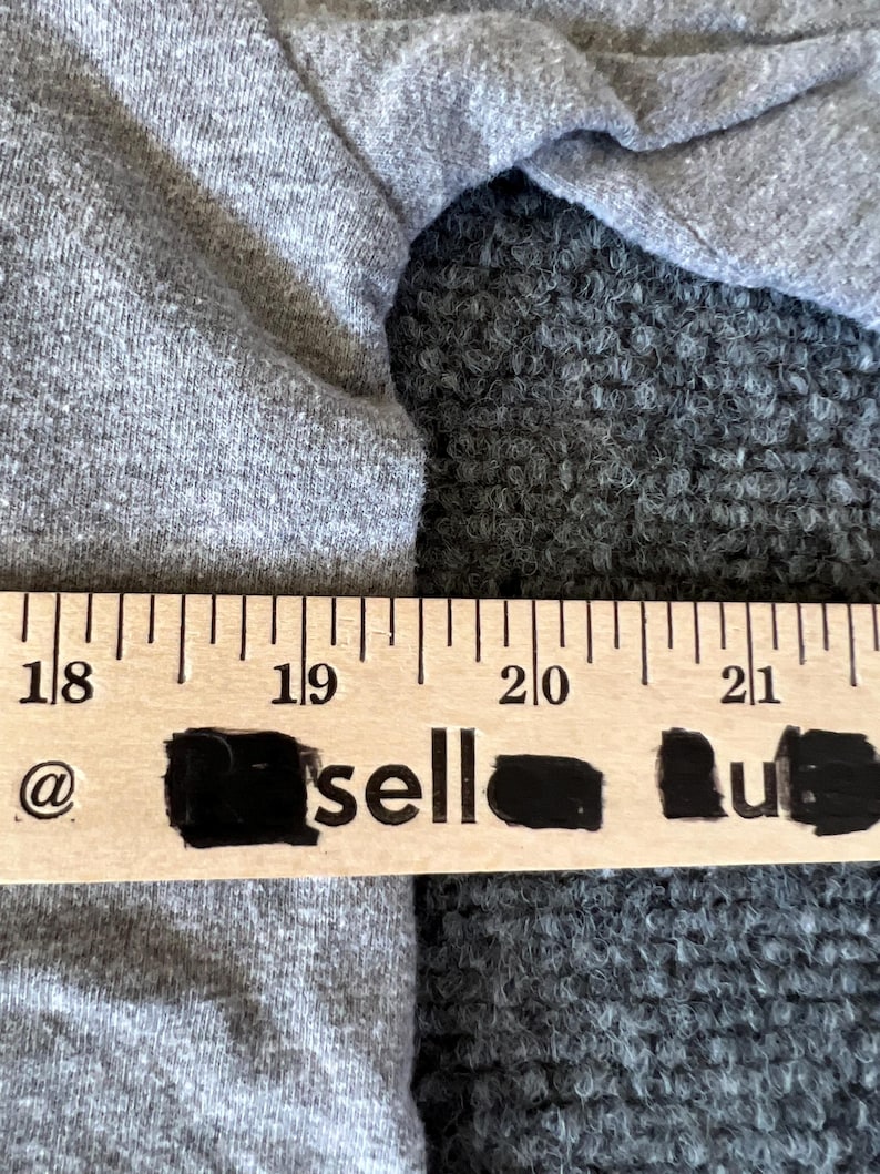 Pu&ograve; includere: Primo piano di un tessuto grigio con un righello di legno sopra. Il righello ha segni neri e la parola "sell" &egrave; parzialmente visibile. Il tessuto sembra essere un materiale a maglia morbido.